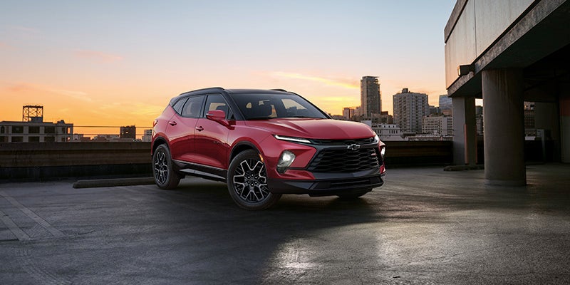 right angle view of a red 2024 chevy blazer parked on a roof open air garage with a sunset and city skyline as a back drop